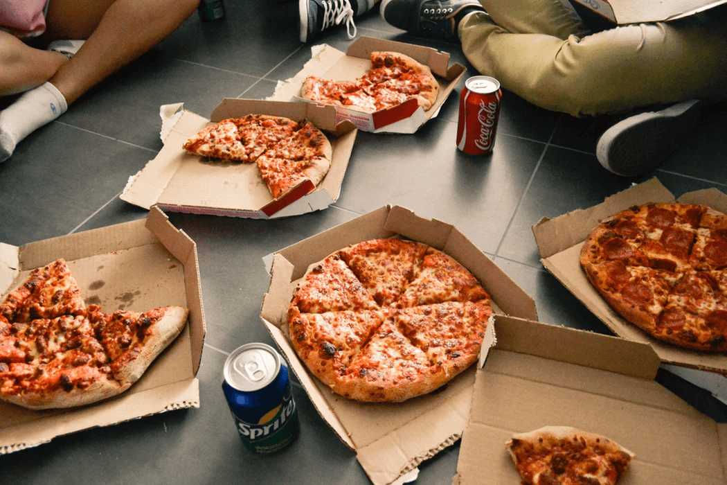 Six open boxes of pizza on the ground with people sitting around them