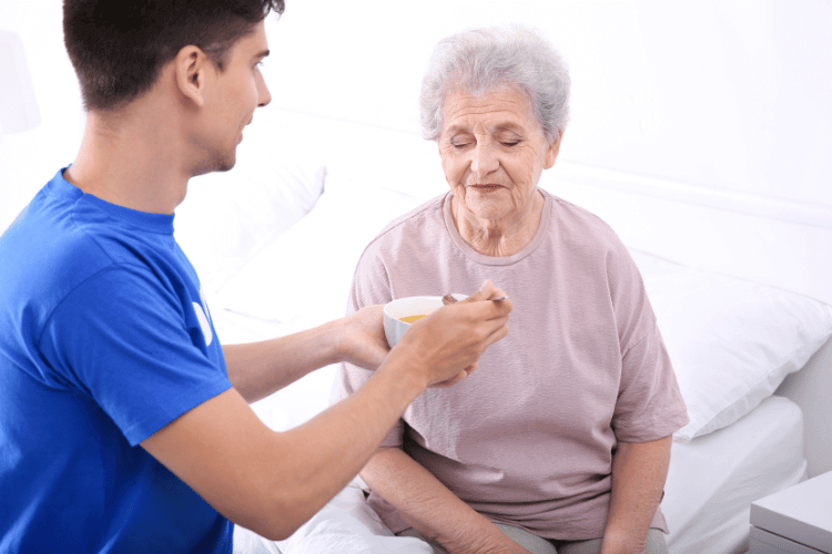 A male caregiver feeding an elderly patient in a bedroom